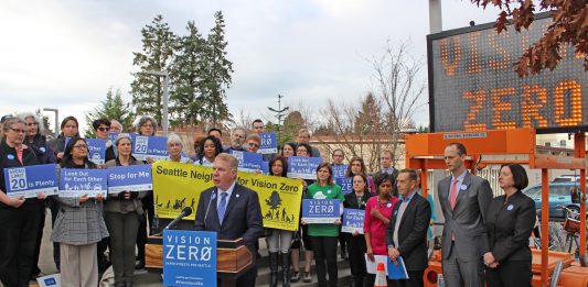 Mayor Ed Murray stands at a podium behind a crowd of staff and advocates with safety-themed signs. An electronic street signs also displays "Vison Zero."