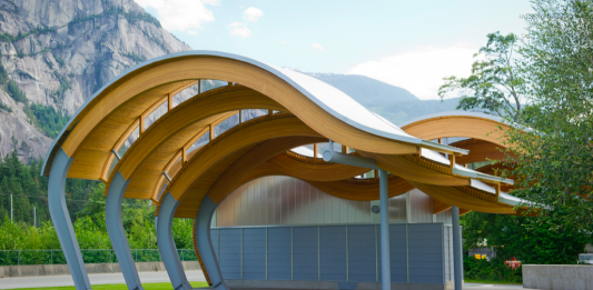 A photo of a wave shaped outdoor pavilion made from wood in front of mountains.