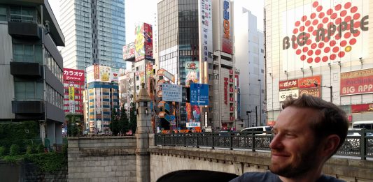 Owen Pickford holding a beer, wearing a Sounders shirt in front of a bridge, river and large towers in Tokyo.