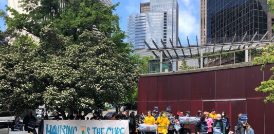 A Stay Housed rally with a "Housing is the Cure" banner at Seattle City Hall with a crowd of about 50.