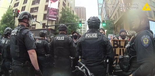 A line of police offers in riot gear face off against protesters sporting Black Lives Matter signs along a Downtown Seattle street.