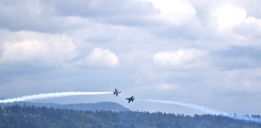 Two Navy Blue Angels planes pass each other over boats tied to a log boom in Lake Washington.