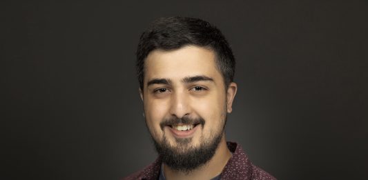 Photo of Rian Watt wearing a t-shirt from the National Civil Rights museum and an unbuttoned button down shirt. He is smiling slightly.