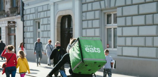 A bike delivery person has a large green box with Uber Eats logo on it strapped to their back while riding down an urban streets with lots of pedestrians.