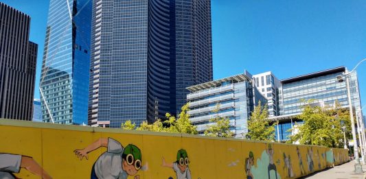 A fence with a pilot boy cartoon figure surrounding Civic Square with Seattle City Hall and the Columbia Tower skyscraper in the background.