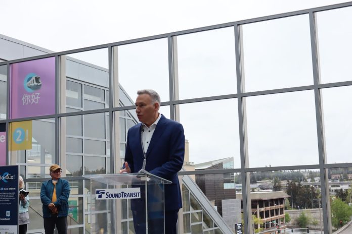 Constantine wears a suit and stands at a glass lectern in from of the Downtown Bellevue Station plaza overlook of the station platform below.