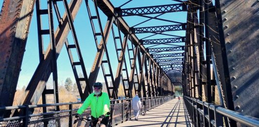 A person bikes on a trestle bridge with a few pedestrians in the background.