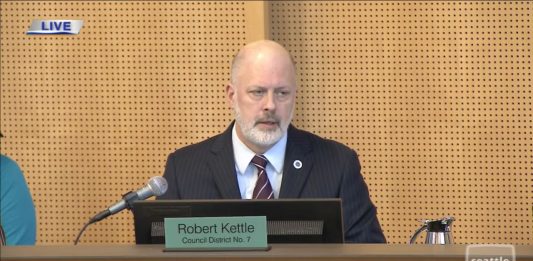 Kattle wears a blue suit and light gray beard and sits on Council dais.