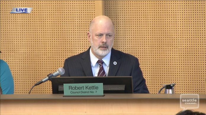 Kattle wears a blue suit and light gray beard and sits on Council dais.