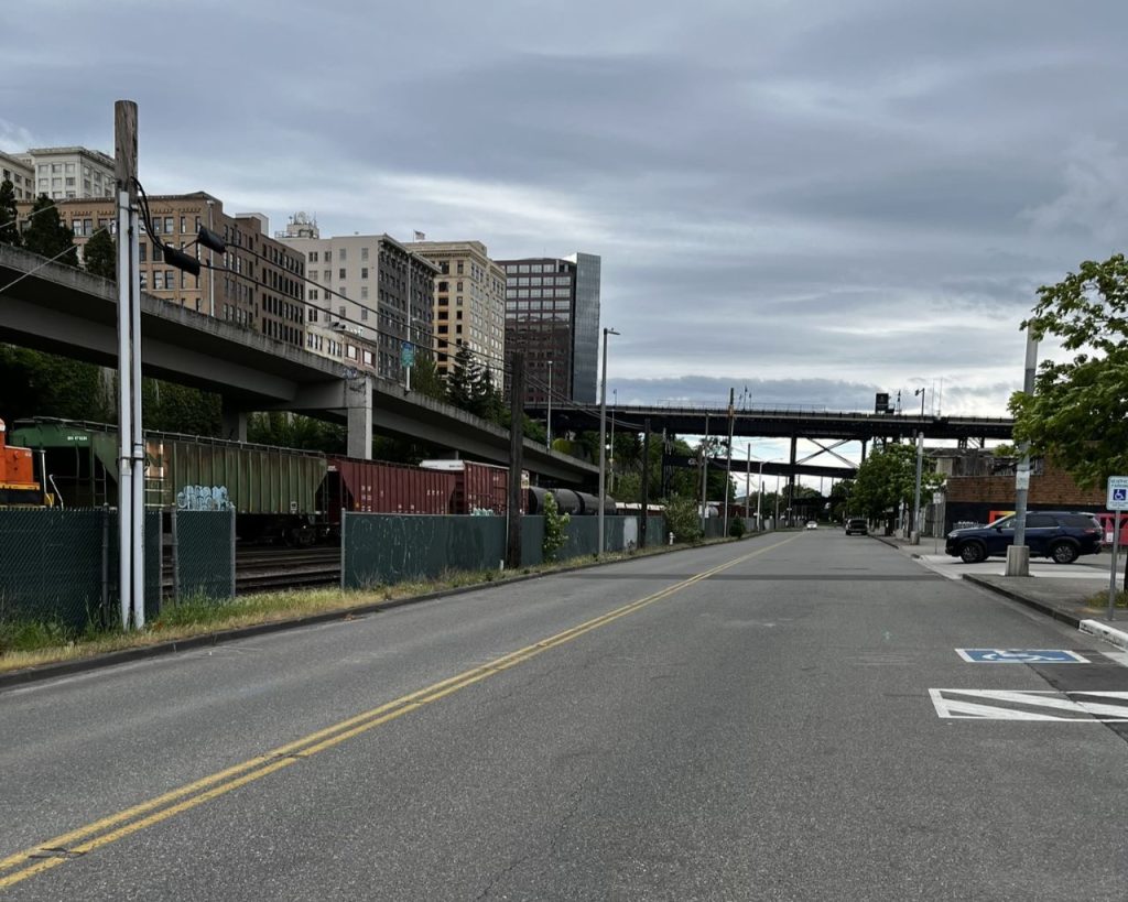 A freight train is in the fenced off railyard with downtown buildings peaking over the ridge. Dock Street is a wide road with no sidewalks on the downtown side.