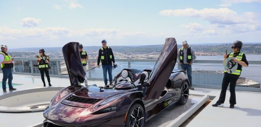 McLaren has its butterfly doors up while a half dozen onlookers in yellow vests take picture or admire the car.