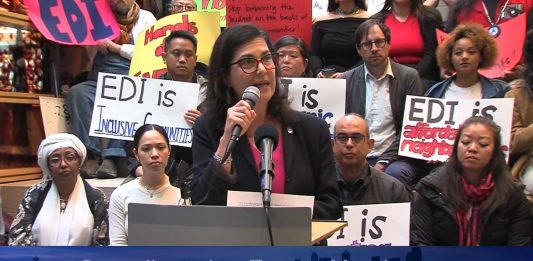 Morales speaks at a podium and several rows of supporters hold signs in support of the EDI program and against the budget raid.