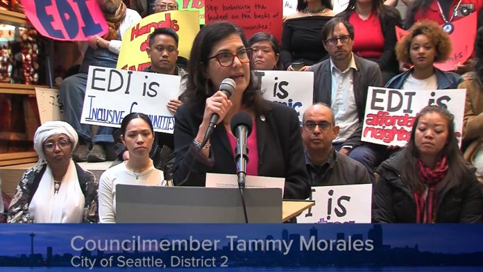 Morales speaks at a podium and several rows of supporters hold signs in support of the EDI program and against the budget raid.