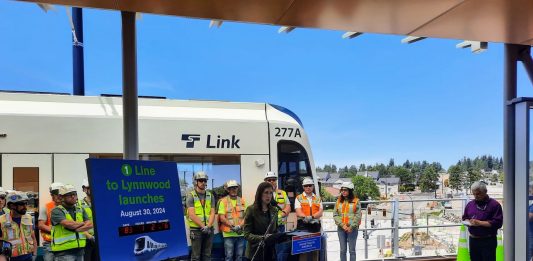 Mestas stands next to a countdown clock with construction workers lined up behind her in front of a Link train.