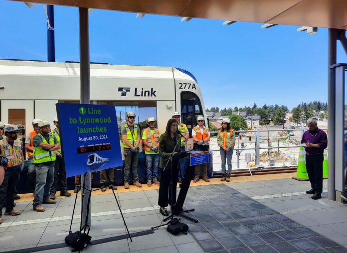Mestas stands next to a countdown clock with construction workers lined up behind her in front of a Link train.