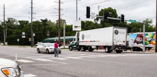 A Black woman crosses in a crosswalk in a very wide busy road as a semi truck zooms by.