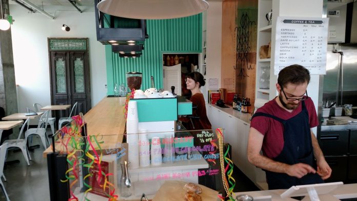 Two servers behind the counter at a cafe, one at the register and another at the espresso machine.