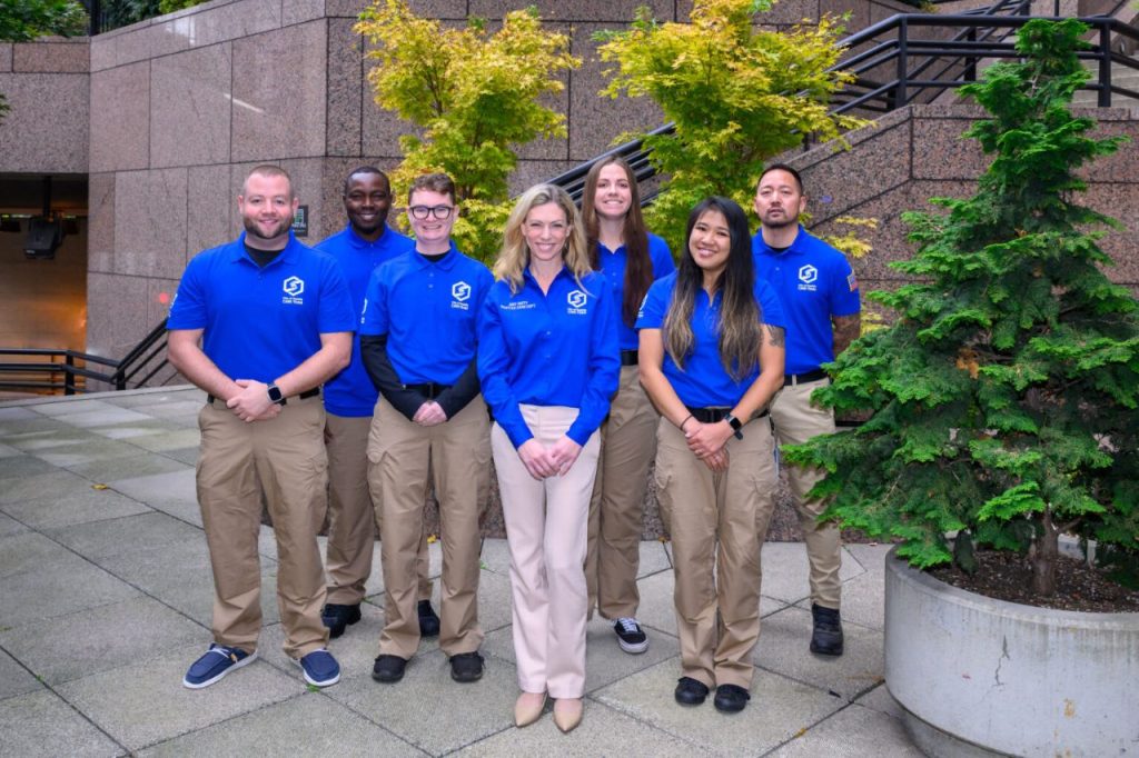 Seven CARE members wear logoed blue short sleeve shirts that are their official garb. These pose of the step of Seattle City Hall.