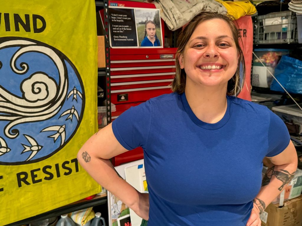 Gemini is a light-skinned woman with tattoos on her arms and a lip ring. She stands next to a tool box and protest flag that says resist and a photo of climate leader Greta Thunberg.