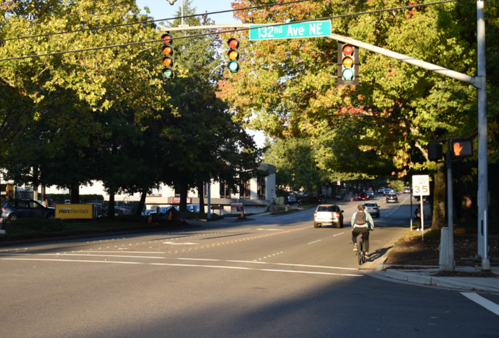 A person biking along the curb of five-lane Bel-Red Road at the intersection with 132th Avenue with cars buzzing by.