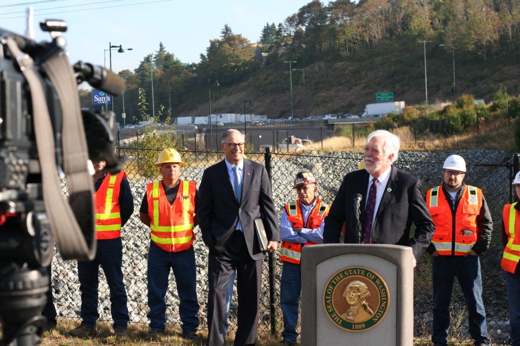 Millar wears a suit and stands at a lectern with Governor Inslee and a group of orange vest wearing construction workers standing behind him.  A busy freeway is in the background.