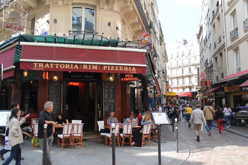 A pedestrianized street with lots of foot traffic next to a sidewalk cafe outside a pizzeria.