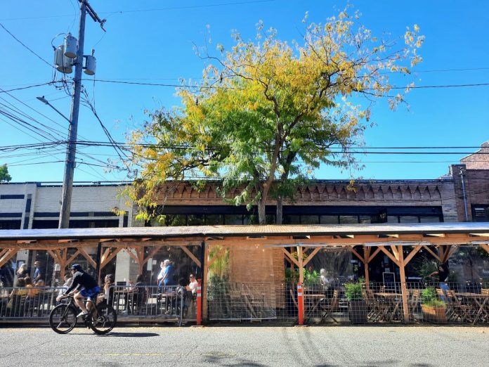 A pergola shelters a street cafe on Ballard Avenue, with a person biking by in the street.