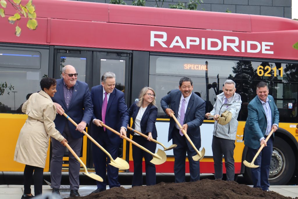 Officials stand in front of a red RapidRide bus with golden shovels with a scoop of ceremonial dirt.