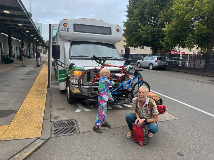 Anna Zivarts and her son smile and load a bike on the rack in front of a bus while it waits at a stop.