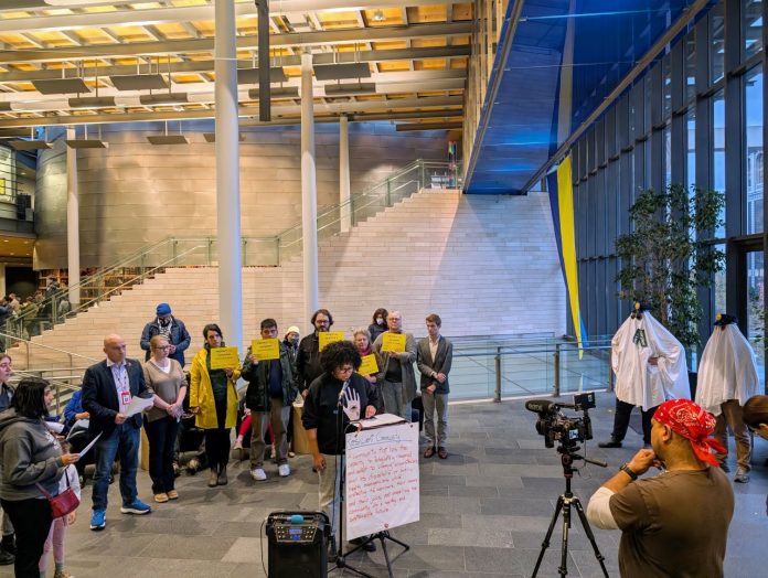 Shemona Moreno is at the lectern with a dozen advocates holding signs in defense of JumpStart investments. Two advocates wear ghost costumes with cop hats representing the ghost cop positions that mayor and council prioritizes for funding.