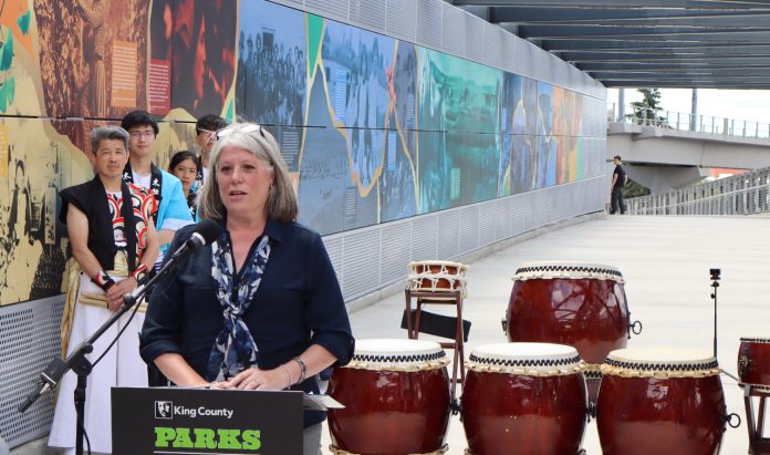 Claudia Balducci stands at a lectern with drums next to her and drummers dressed in traditional Japanese garb behind.