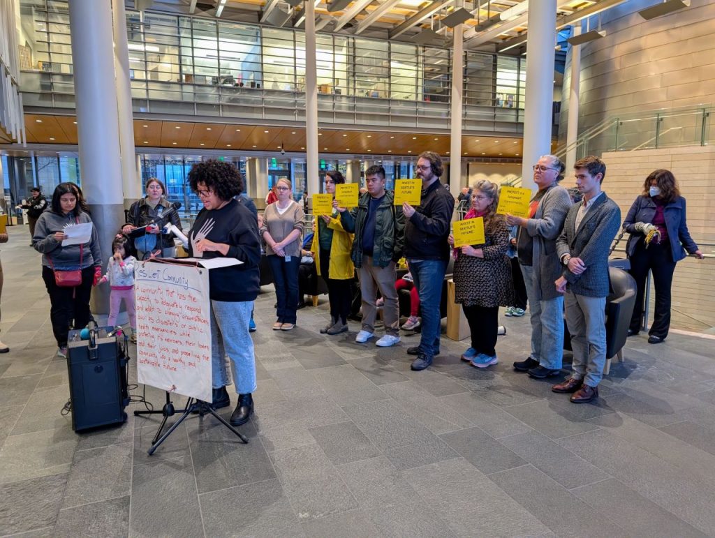 A sign reads Resilient communities on the lectern in the lobby of city hall and people stand behind the speaker holding signs saying a protest JumpStart commitments and invest on Seattle's future.