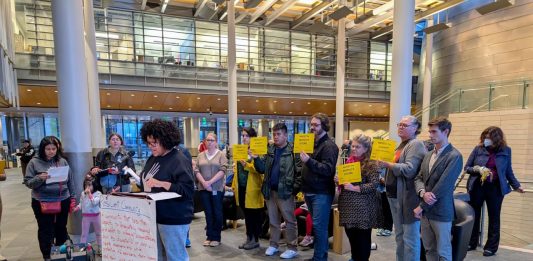 A sign reads Resilient communities on the lectern in the lobby of city hall and people stand behind the speaker holding signs saying a protest JumpStart commitments and invest on Seattle's future.