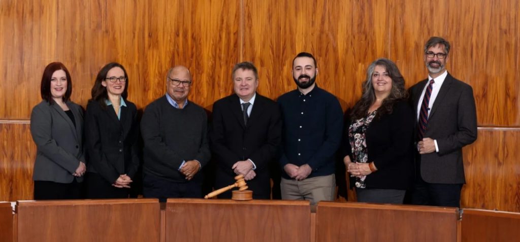 Members stand in the wood-panel council chamber behind a desk with a gavel.