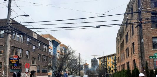 Pedestrians cross Denny Way at the Harvard Avenue intersection with midrise apartment buildings in the background.