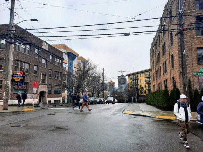 Pedestrians cross Denny Way at the Harvard Avenue intersection with midrise apartment buildings in the background.