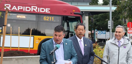 Greg Spotts standing at a podium with Bruce Harrell looming over and a RapidRide bus behind