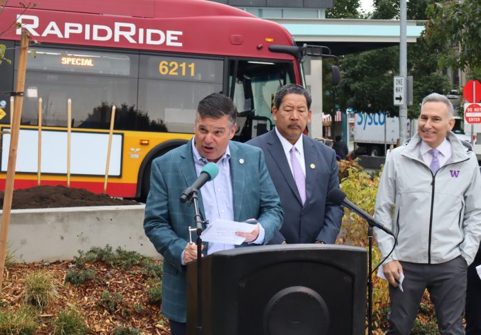 Greg Spotts standing at a podium with Bruce Harrell looming over and a RapidRide bus behind