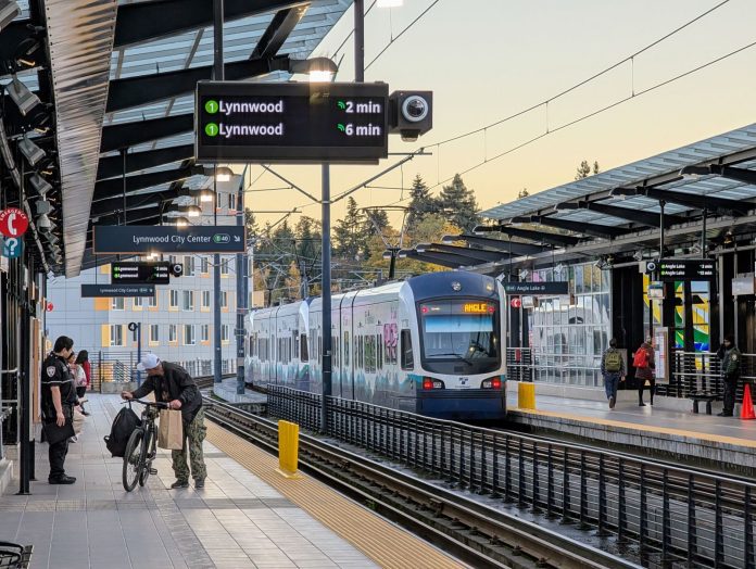 A southbound train departs with an apartment building in the background. A man rolls a bike on the platform and security guards stand around.