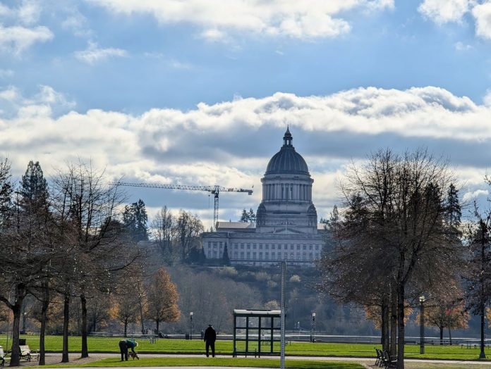 The domed Olympia Capitol Building stands in the distance with a bus stop with a passenger waiting in the foreground. A dogwalker also mosey on the lawn.