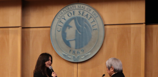 The pair stand before the Seattle city seal at Seattle City Council chambers.