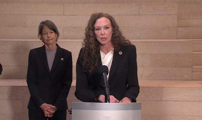 Davison is a white woman with curly hair in a dark blazer. She stands at a lectern in city hall with Sara Nelson behind her.