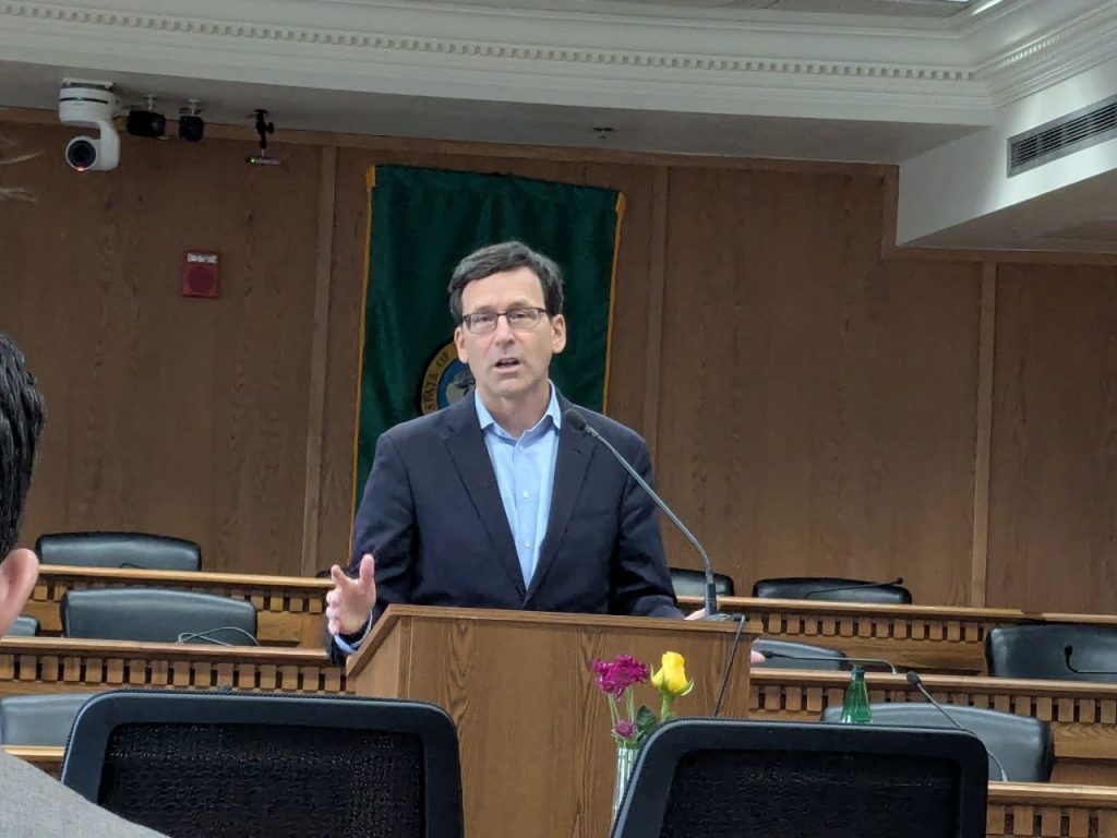 Ferguson stands at the lectern wearing a navy suit and glasses in a Capitol board room.