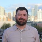 A bearded man smiles on a rooftop with the Seattle skyline in the background.