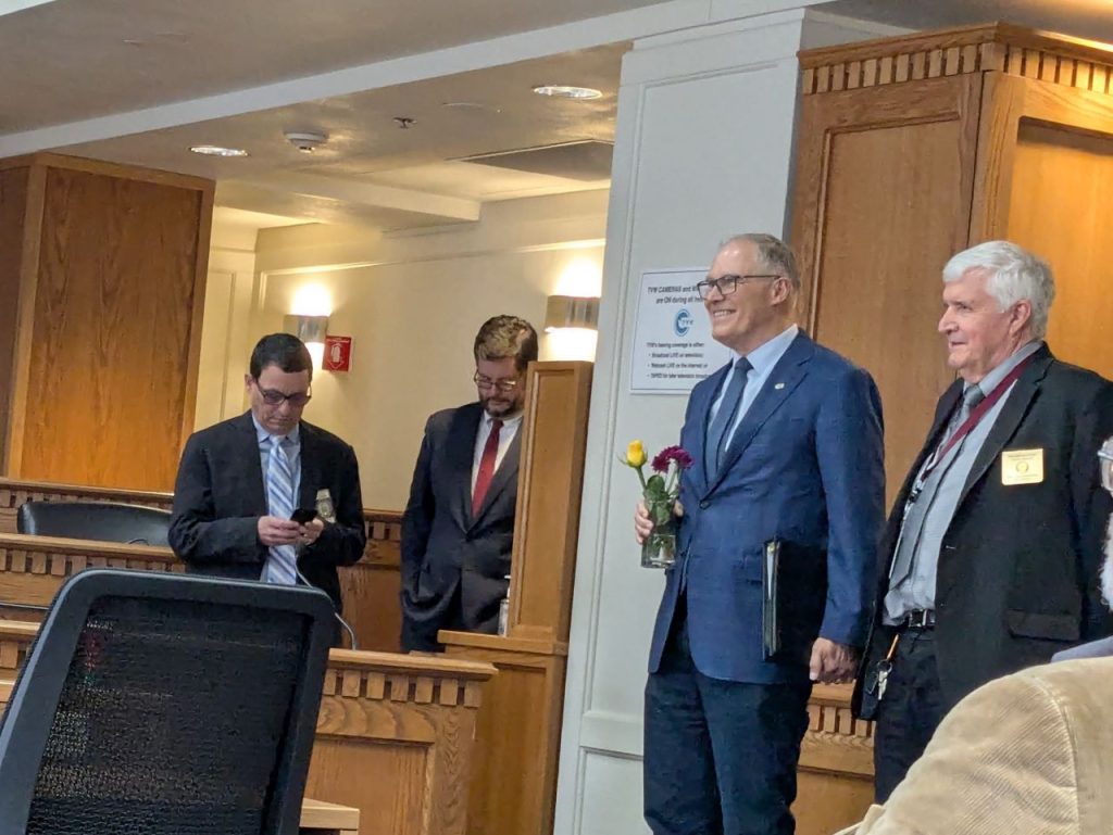 Inslee holds a small vase of flowers and holds hands with a staff member as if they are about to walk down the altar.