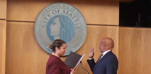 Solomon is a bald black man wearing eyeglasses and a dark suit. He stands in front of the Seattle City seal with his hand raised for his swearing in.