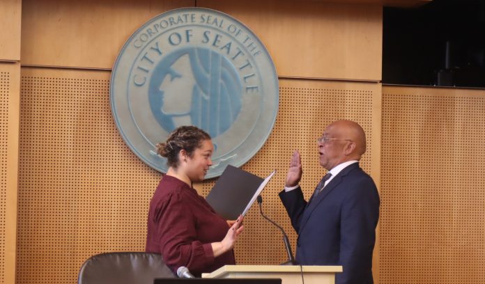 Solomon is a bald black man wearing eyeglasses and a dark suit. He stands in front of the Seattle City seal with his hand raised for his swearing in.