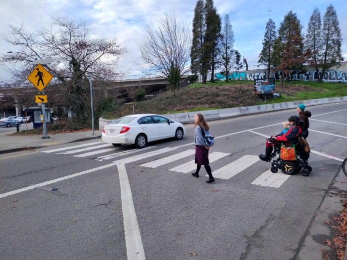 Two people crossing the ramp with one car already having disregarded them. One person is in a wheelchair.