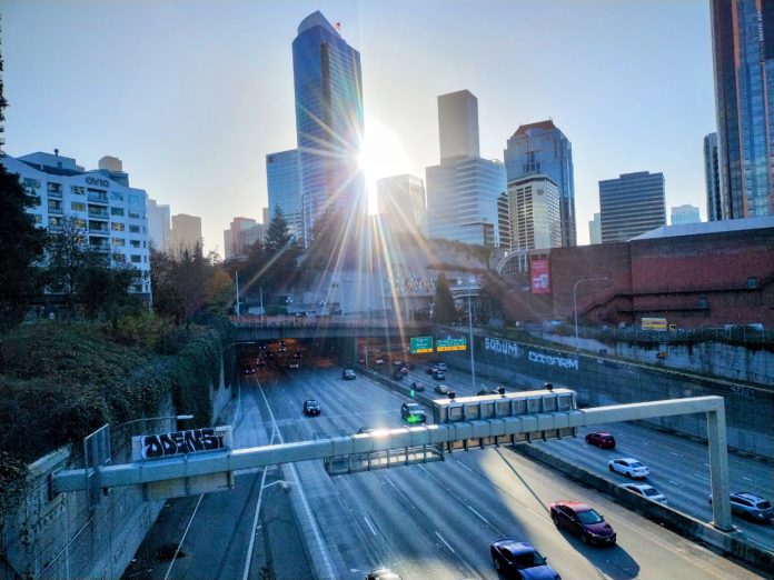 Cars use Interstate 5 through Seattle on a sunny day, with the sun just breaking through the city skyline