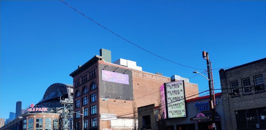 A photo from SoDo, with T-Mobile Park in the background. An older brick building stands next to smaller, more run down storefronts with a large wide road in the foreground